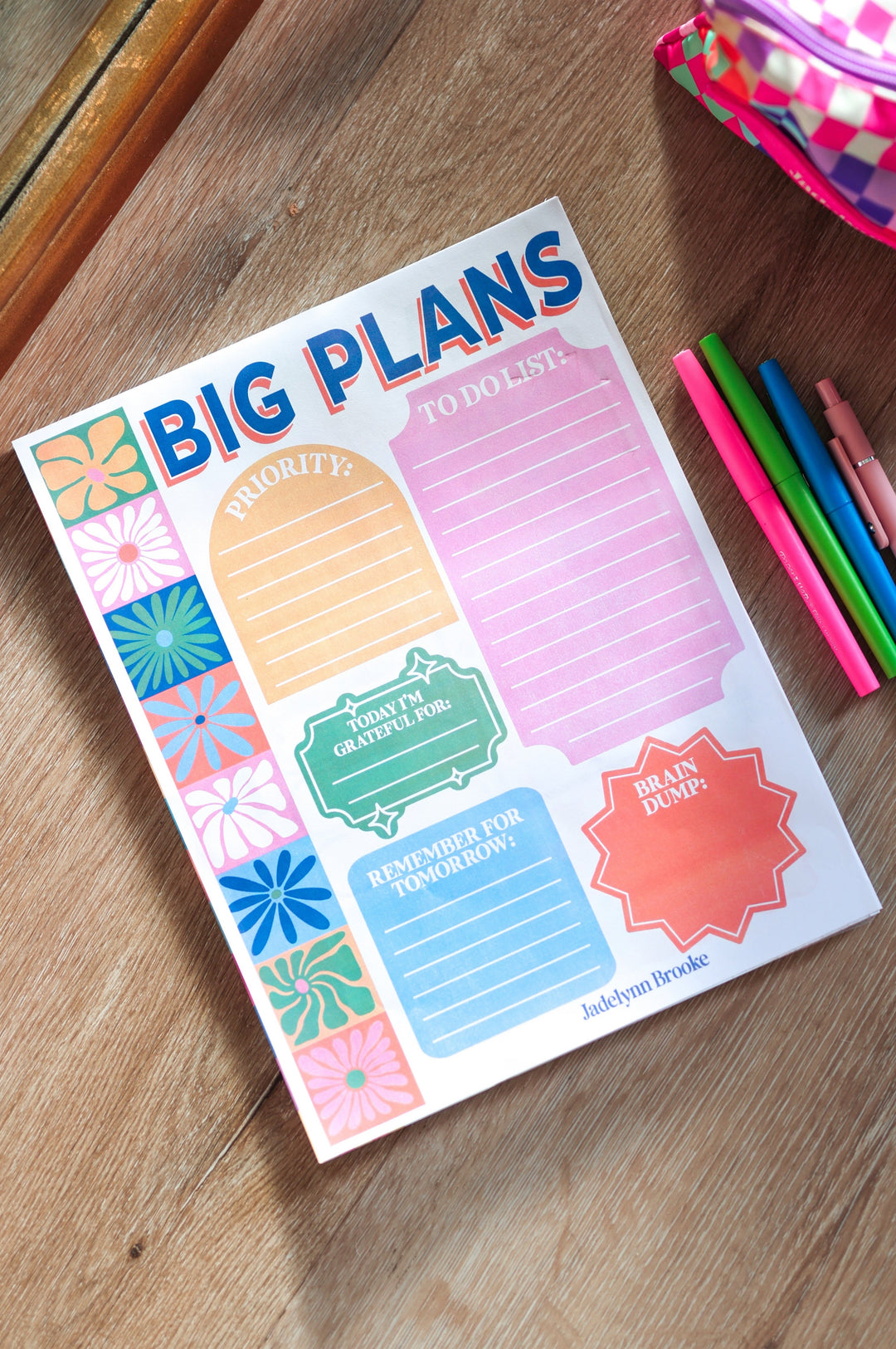 Colorful notepad with 'BIG PLANS' on a wooden surface with pens and a pink pouch.