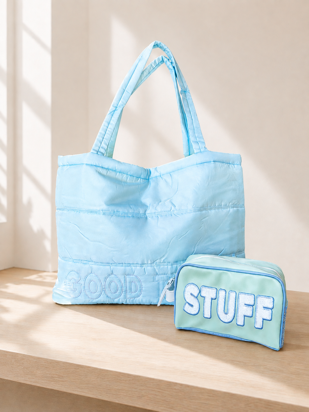 A light blue quilted tote bag with 'GOOD TIMES' embroidered on the bottom, displayed next to a matching light green zippered pouch labeled 'STUFF' on a wooden shelf against a beige wall with soft window shadows.