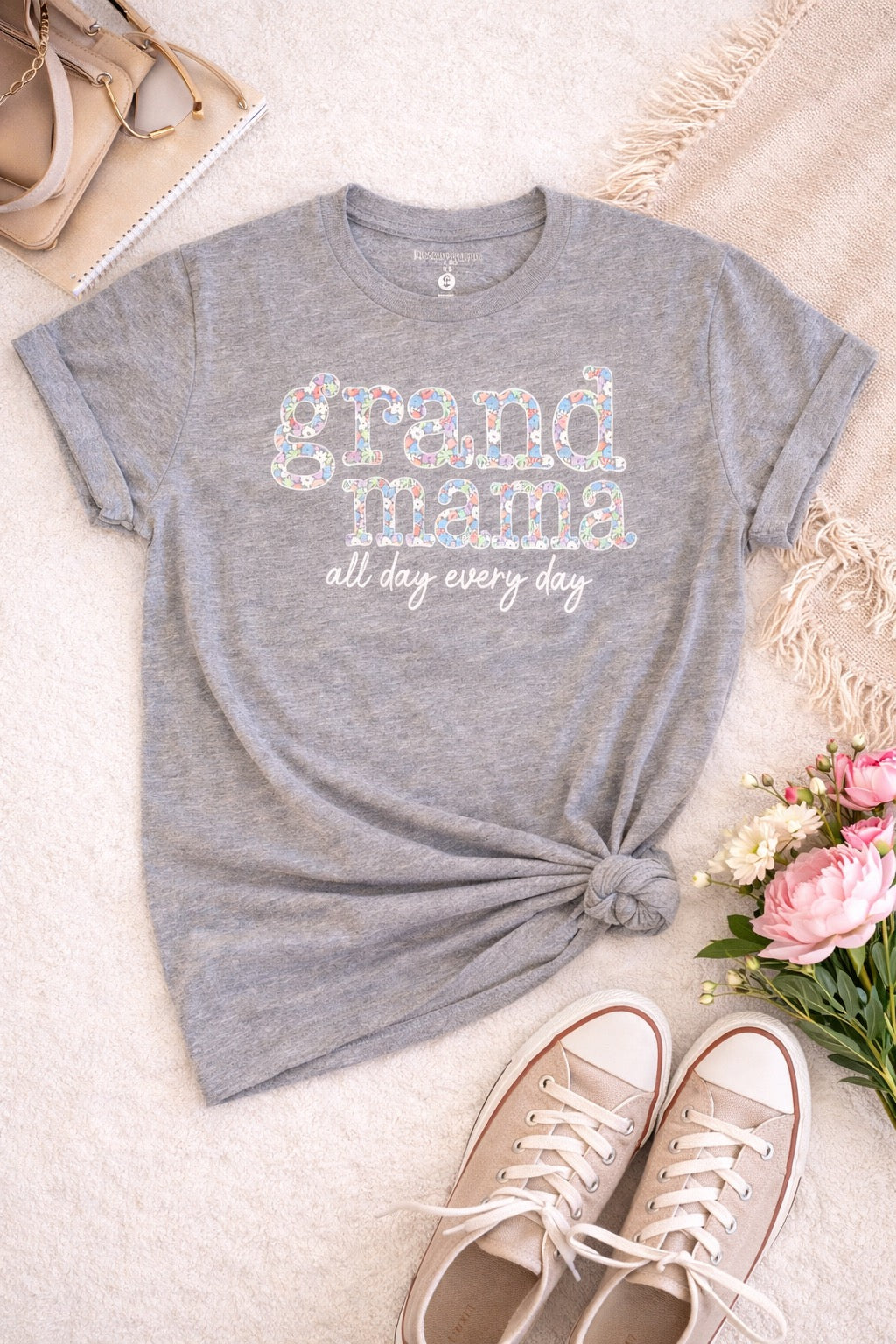 Gray t-shirt with 'grand mama all day every day' text, paired with shoes and flowers on a light surface.