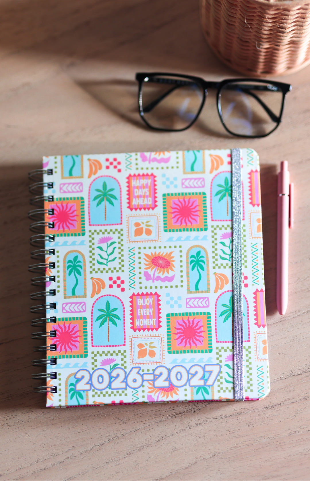 Colorful planner with 2026-2027 text on a wooden surface with glasses and pen.