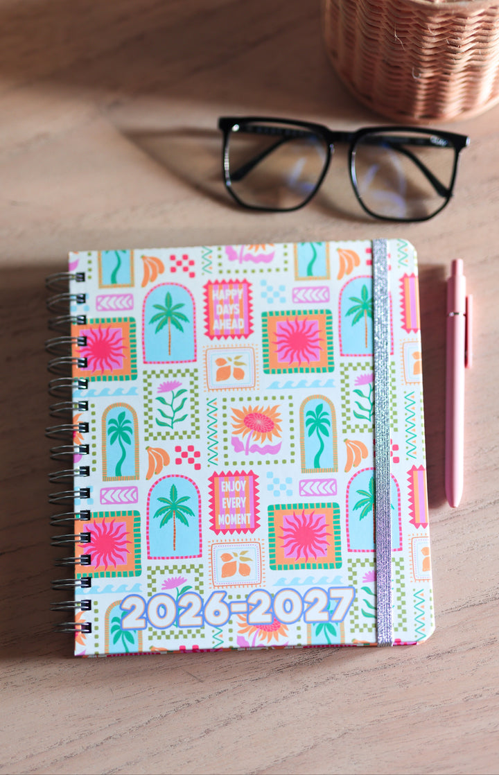 Colorful planner with 2026-2027 text on a wooden surface with glasses and pen.