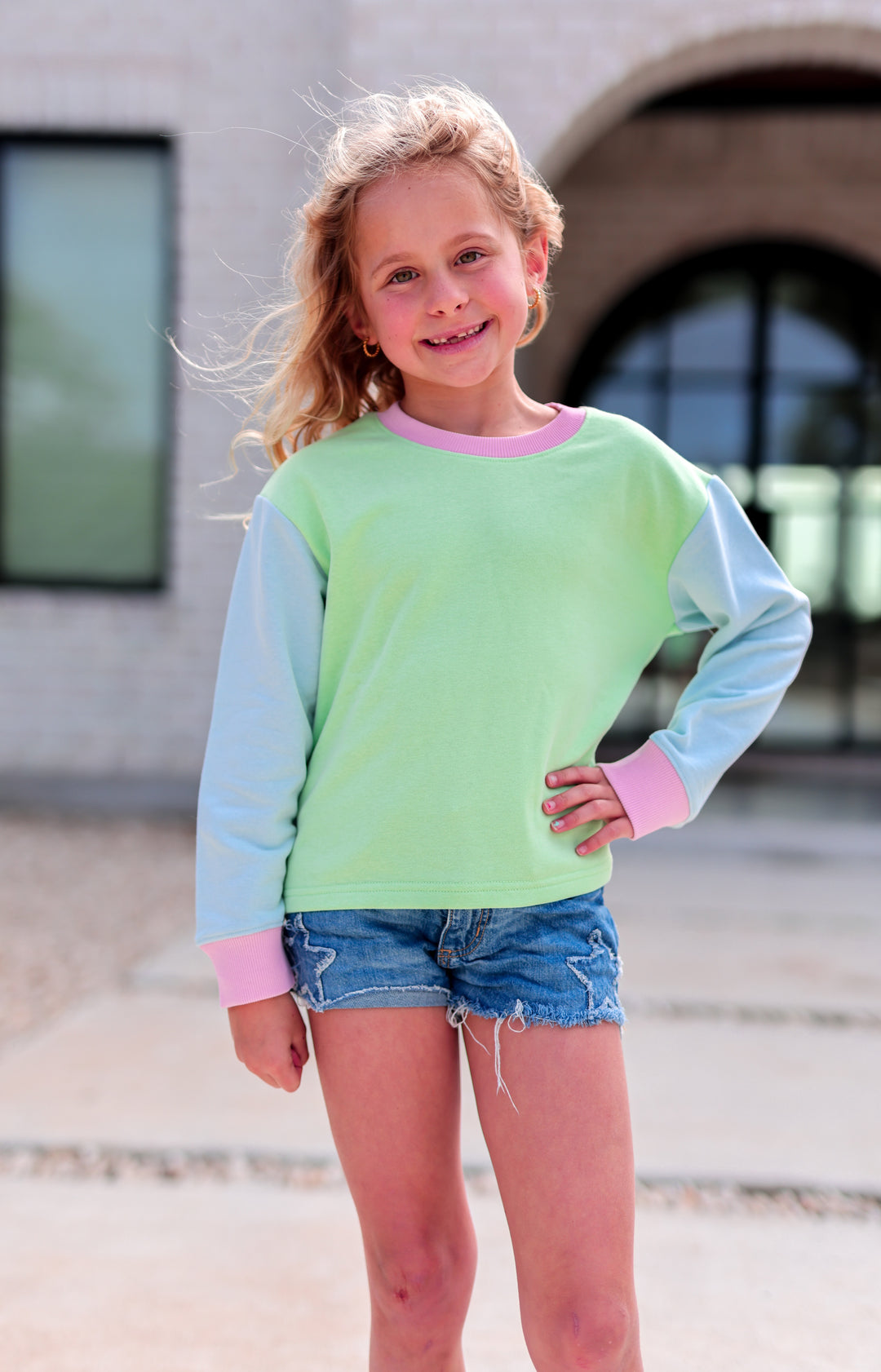 Girl wearing a pastel green and blue long-sleeve sweatshirt, styled with denim shorts.