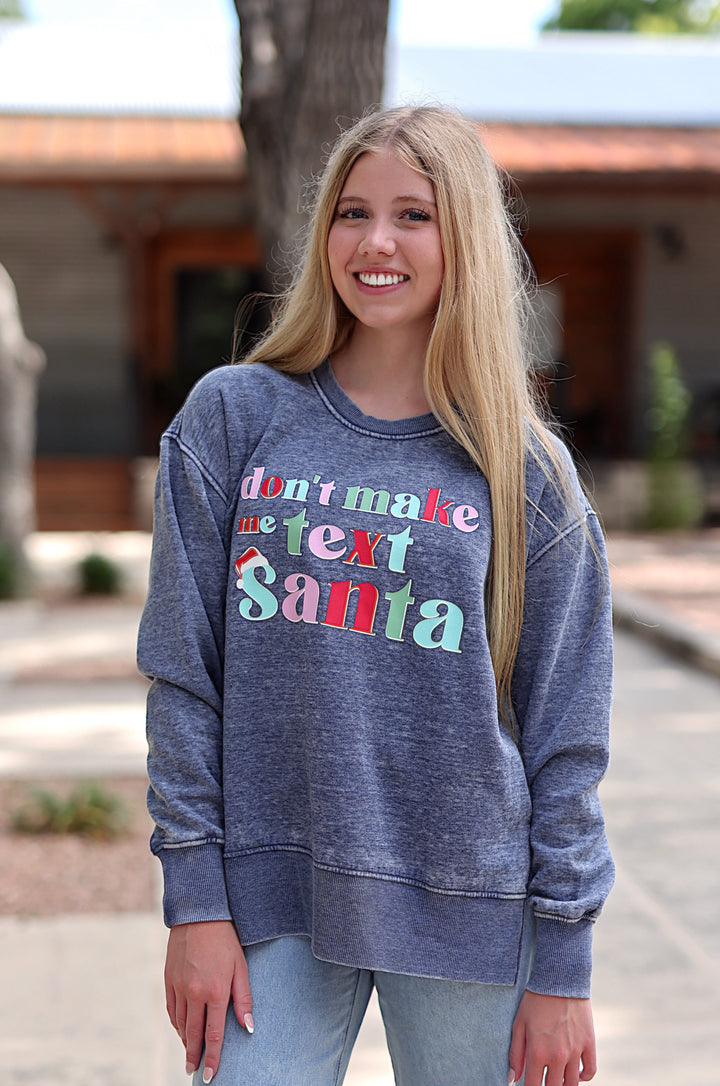 A smiling young woman stands outdoors on a sidewalk wearing a faded heathered blue-gray crewneck sweatshirt with a colorful graphic that reads 'don't make Santa a pest' and light-wash jeans.