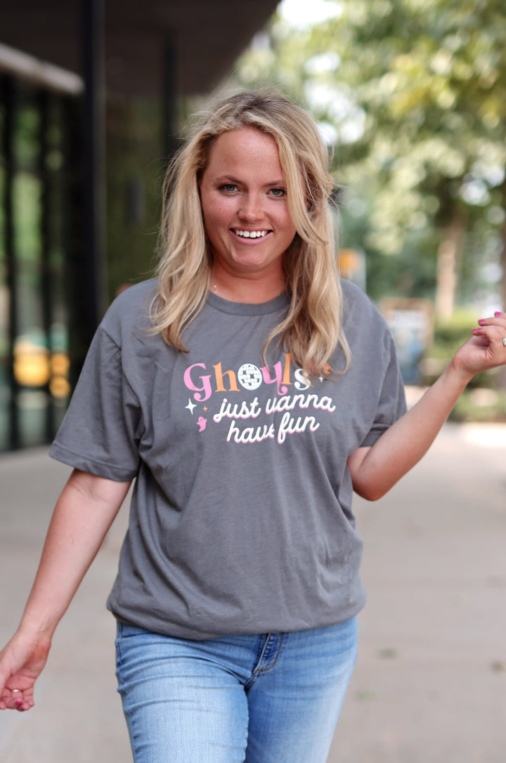 A woman walking outdoors on a sidewalk wearing a heather gray graphic t-shirt with the text 'Ghouls just wanna have fun' in pink and white, paired with light blue distressed jeans.