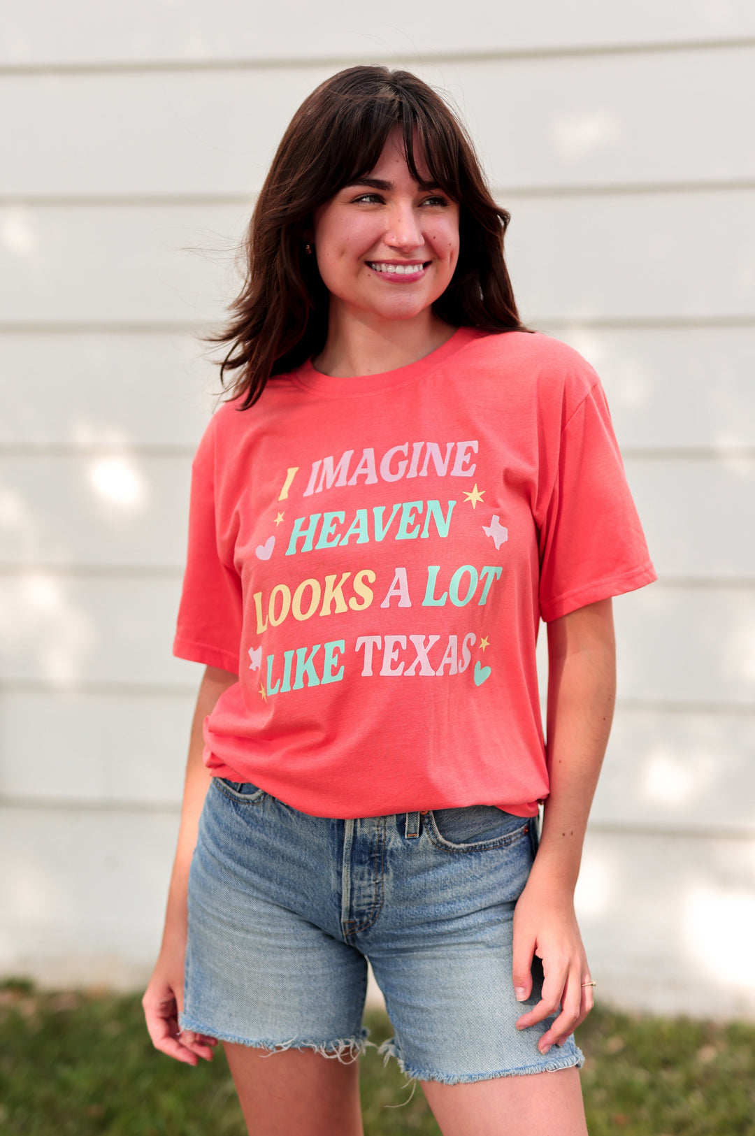 Woman wearing a orange t-shirt with text, standing outdoors against a white wall.