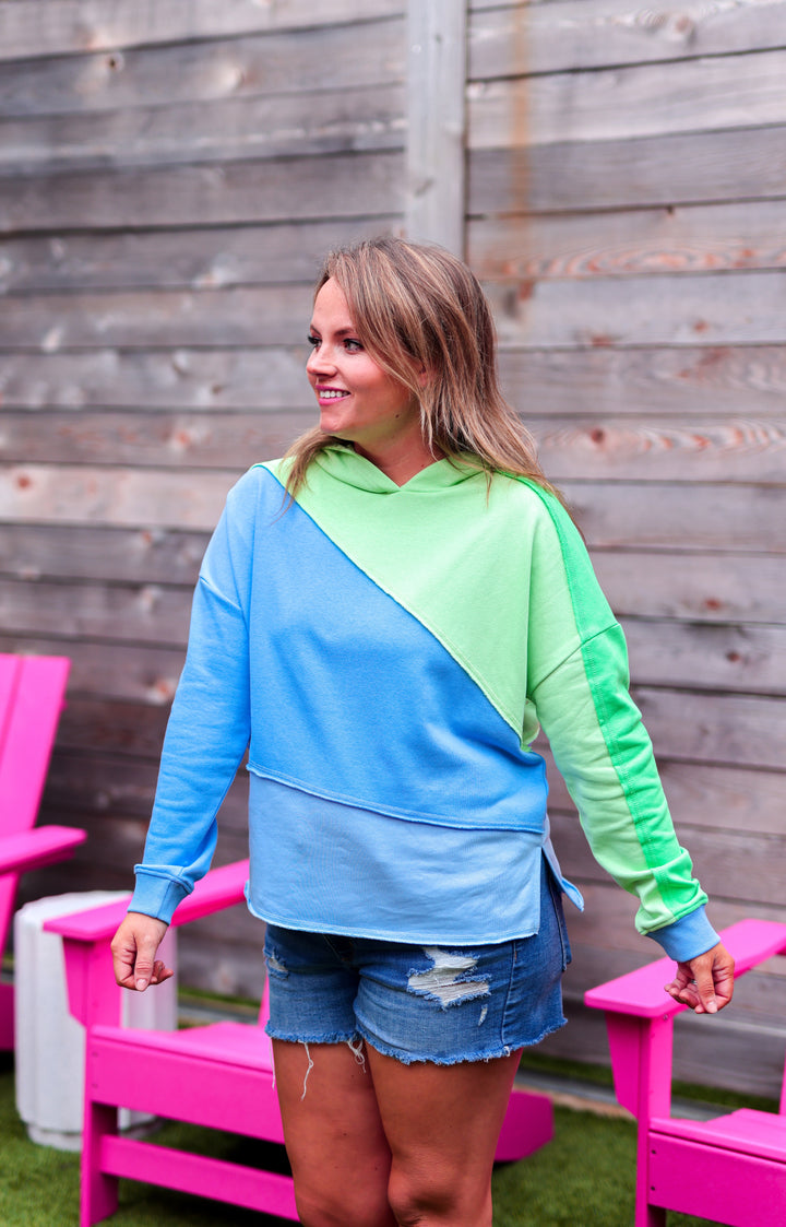 Woman wearing a blue and green color-blocked hoodie sitting on pink chairs outdoors.