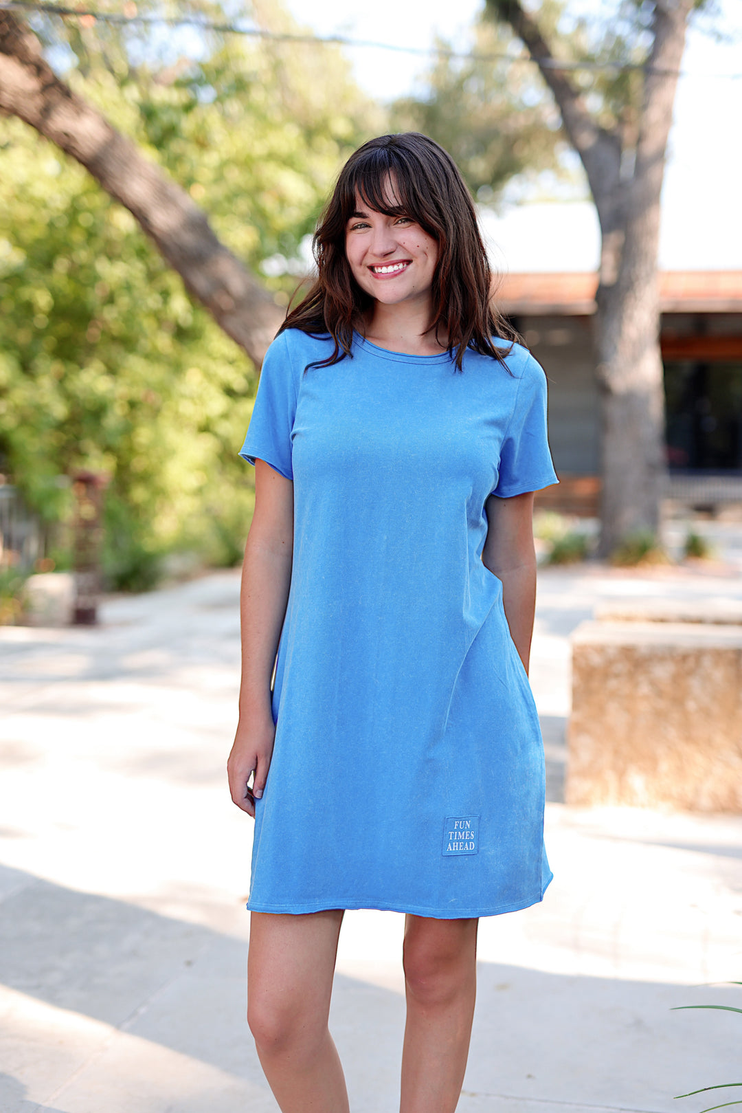 Woman wearing a blue dress standing outdoors with trees and a building in the background