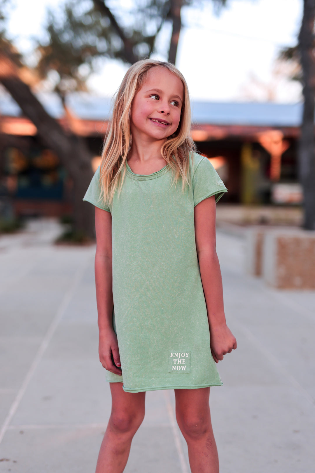 Young girl wearing a green dress with text, standing outdoors.