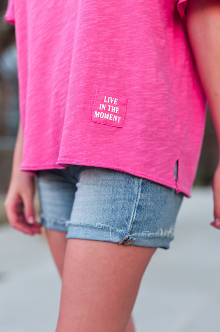 Women's pink t-shirt with 'Live in the Moment' patch, paired with denim shorts for a casual summer look.