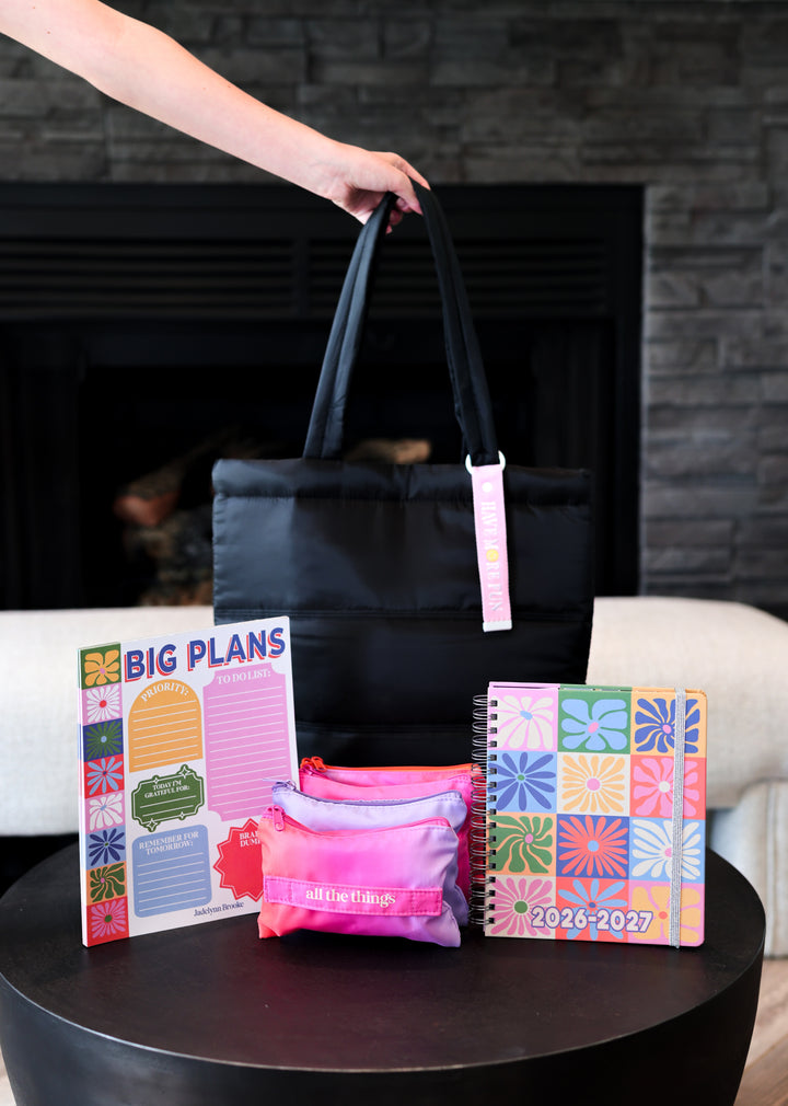 Black tote bag with pink accents on a table next to planners and a notebook.