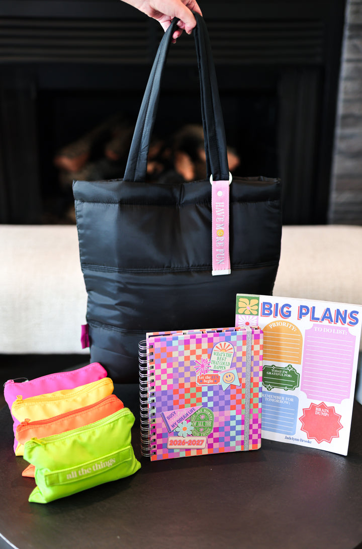 Black tote bag with colorful items including a planner and pouches on a table.