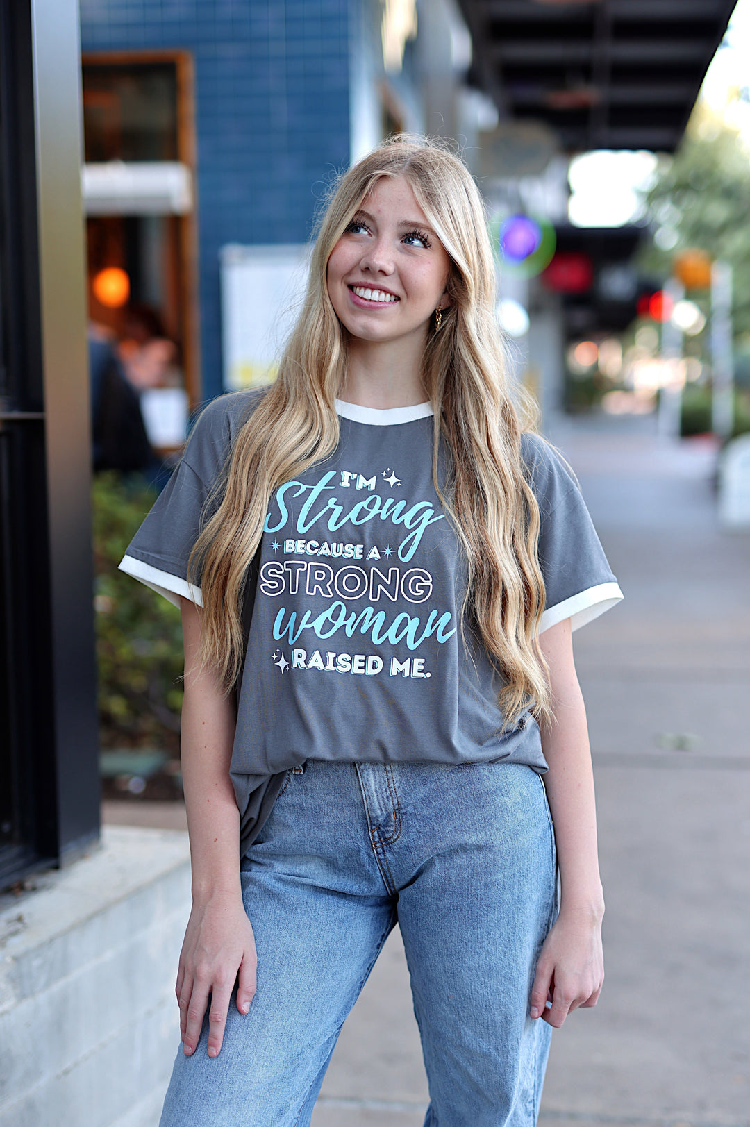 Woman wearing a gray t-shirt with a motivational quote, standing on a sidewalk.