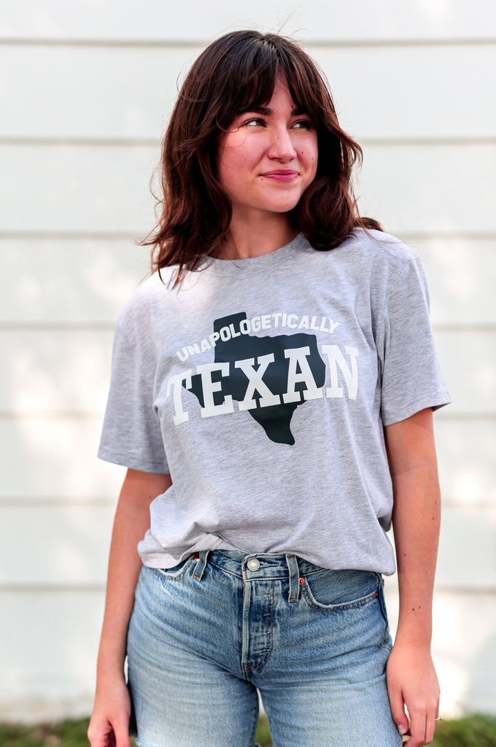 Person wearing a gray t-shirt with 'Unapologetically Texan' text and denim shorts outdoors.