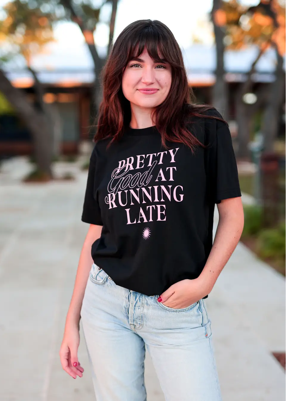 Woman wearing a black t-shirt with 'Pretty Good at Running Late' text, standing outdoors.