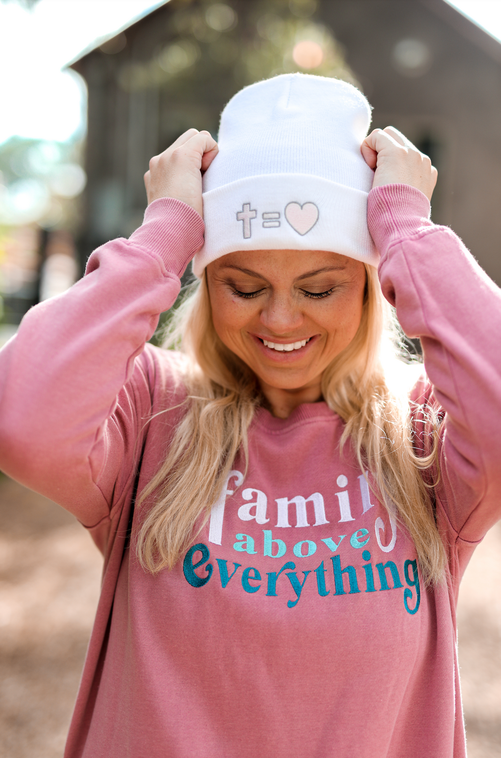 Woman smiling in a pink sweatshirt and white beanie with cross and heart design, outdoors.